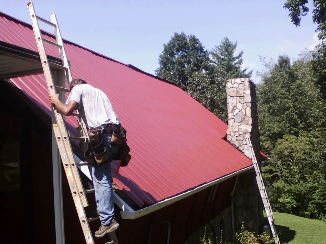 Roofer on a ladder beside a red metal roof and stone chimney — Soffit & Fascia Repair by Roofing Brothers in Saint Catharine
